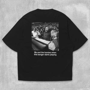 May include: Black t-shirt with a black and white image of people reacting excitedly. The text "Me and the homies when this banger starts playing" is below the image. The shirt is made of a soft material.