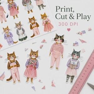 K&ouml;nnte beinhalten: Ein Blatt Papier mit Katzenillustrationen und dem Text Print, Cut & Play 300 DPI, zusammen mit mehreren einzelnen Katzen-Papierpuppen-Ausschnitten in verschiedenen Outfits und einem rosa Lineal mit einer L&auml;nge von etwa 20 cm.