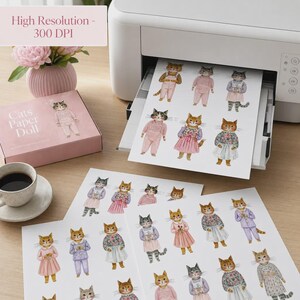 K&ouml;nnte beinhalten: Ein wei&szlig;er Drucker gibt ein Blatt Papier mit mehreren Aquarellillustrationen von Katzen in verschiedenen Outfits aus, neben einer rosa Schachtel mit der Aufschrift Cats Paper Doll und einer Vase mit rosa Blumen. Das Papier zeigt Katzen in Kleidern.