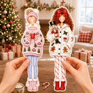 May include: Two die-cut paper dolls featuring festive winter attire. The left doll wears a pink sweater with house and tree patterns, holding a red mug. The right doll has red curly hair, a white cardigan with holiday motifs, and holds a nutcracker.