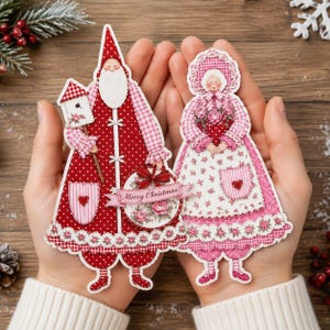 Pode incluir: Dois ornamentos de papel recortado com uma figura de Pai Natal num casaco vermelho às bolinhas e uma figura de Mãe Natal num vestido floral rosa. O Pai Natal segura uma casa de pássaros e um sinal Merry Christmas, ambos com renda.