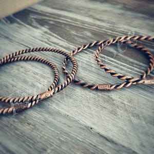 May include: Two circular bangles crafted from twisted copper wire. Each bangle features a small, cylindrical metal connector piece embossed with a subtle, swirling scroll pattern. The copper has a warm, metallic finish with dark oxidation.