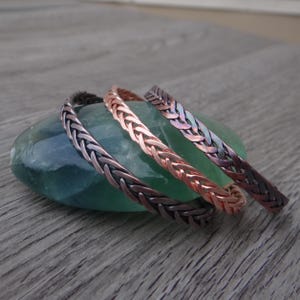 May include: Three metal bracelets featuring a woven, braided pattern. The bracelets are displayed in varying metallic tones, including a dark oxidized finish, a bright copper color, and a multi-colored iridescent finish with purple and blue hues.