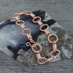 May include: A copper bracelet featuring a repeating pattern of twisted wire circular links connected by segments of tightly coiled copper wire. The bracelet includes a simple hook-and-eye style clasp mechanism made from the same copper wire material.