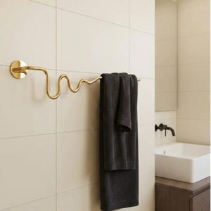 May include: A gold-toned metal towel rail with a wavy, serpentine design mounted on a tiled wall. A dark grey, ribbed-texture towel is draped over the rail. The towel rail features a circular wall mount on the left side and a curved, undulating rod.