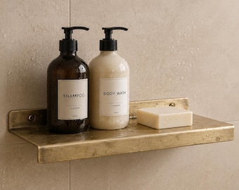 May include: A brass-toned metal shelf holding a dark amber bottle labelled SHAMPOO, a light beige bottle labelled BODY WASH, and a rectangular off-white bar of soap. The shelf is mounted horizontally and features a simple, flat design.