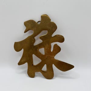 May include: A brass-colored metal wall hanging featuring an intricate, cutout design of a Chinese character. The object has a smooth, reflective surface with a warm, metallic tone and irregular, organic edges that form a stylized, calligraphic shape.