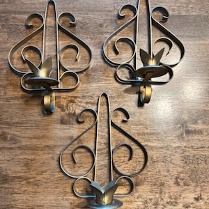 May include: Three gold-toned metal wall sconces featuring a symmetrical scrollwork design. Each sconce has a central vertical bar, curved wire accents, and a small circular platform at the base designed to hold a single candle securely in place.