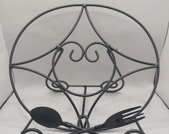 Op de afbeelding: Een donkergrijze metalen displaystandaard met een cirkelvormig frame en een centraal hartvormig ontwerp. De basis wordt ondersteund door twee krulvoeten en is aan de voorzijde versierd met een gekruist lepel- en vorkmotief.