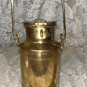 May include: A cylindrical brass milk can with a hinged lid, a latch, and a tall arched handle. A yellow measuring tape is positioned at the base, showing the container has a diameter of approximately 6 inches at its widest point.