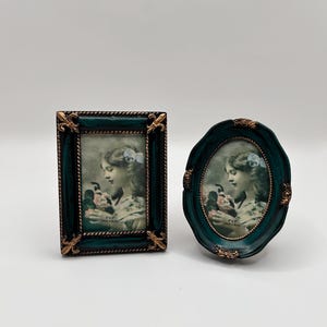 May include: Two dark teal picture frames with gold-toned fleur-de-lis accents. One frame is rectangular and the other is oval. Both contain a vintage-style photograph of a person holding flowers, with text indicating a 2x3 inch photo size.