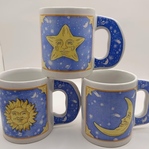 May include: Three white ceramic mugs featuring celestial designs. Each mug displays a blue background with yellow illustrations of a sun, a moon, or a star, all with facial features. The handles are also decorated with blue and white star patterns.