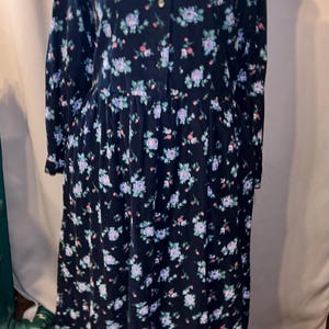 May include: A long-sleeved, dark navy blue dress featuring a repeating floral pattern in shades of light blue, pink, and green. The dress has a collared neckline, a full button-down front closure, and a gathered waistline with a full skirt.