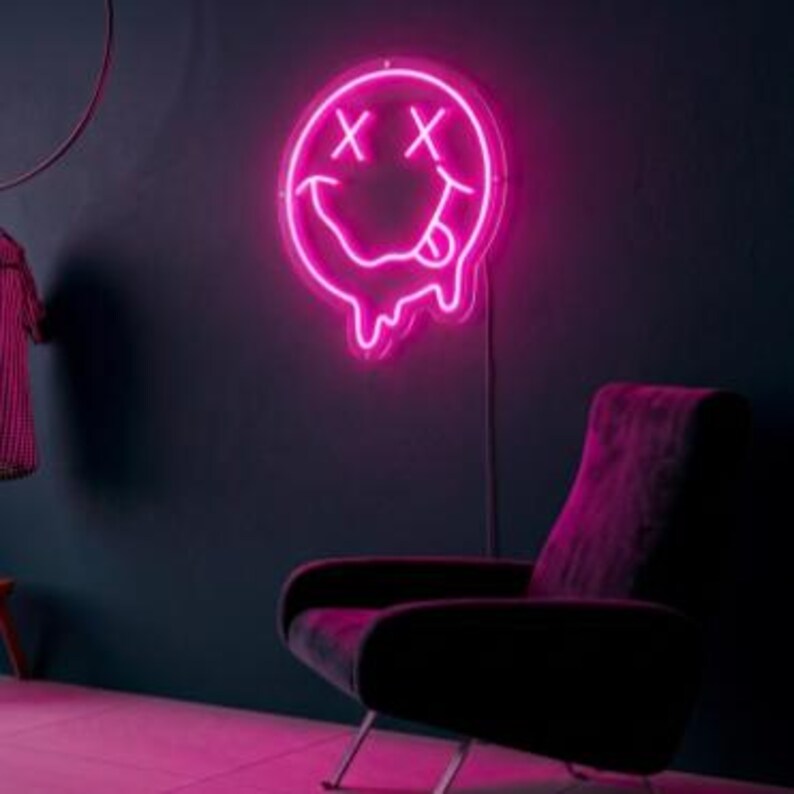 May include: A bright pink neon sign shaped like a circular smiley face with X-shaped eyes and a tongue sticking out, featuring a melting effect at the bottom edge. The neon light is mounted on a dark wall above a dark-colored upholstered armchair.