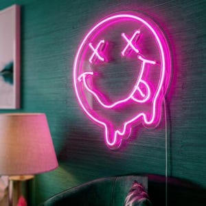May include: A bright pink neon sign shaped like a circular smiley face with X-shaped eyes and a tongue sticking out. The bottom of the circular face features a melting effect with dripping neon lines, mounted on a clear backing with a power cord.