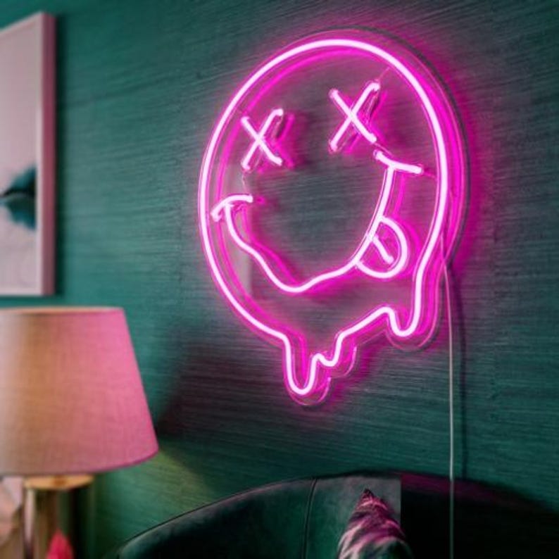 May include: A bright pink neon sign shaped like a circular smiley face with X-shaped eyes and a tongue sticking out. The bottom of the circular face features a melting effect with dripping neon lines, mounted on a clear backing with a power cord.