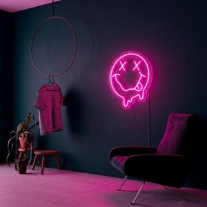 May include: A bright pink neon sign featuring a melting smiley face with X-shaped eyes and a tongue sticking out. The sign is mounted on a dark wall above a dark armchair. A checkered shirt hangs from a circular rack nearby.