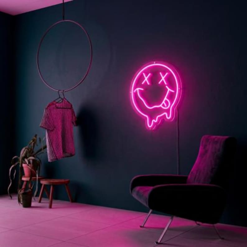 May include: A bright pink neon sign featuring a melting smiley face with X-shaped eyes and a tongue sticking out. The sign is mounted on a dark wall above a dark armchair. A checkered shirt hangs from a circular rack nearby.
