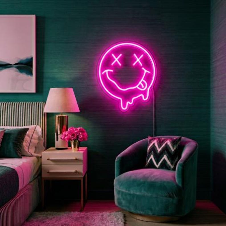 May include: A bright pink neon sign shaped like a melting smiley face with X-shaped eyes and a tongue sticking out. The circular light fixture features a glowing outline and a dripping effect at the bottom, mounted on a dark green textured wall.