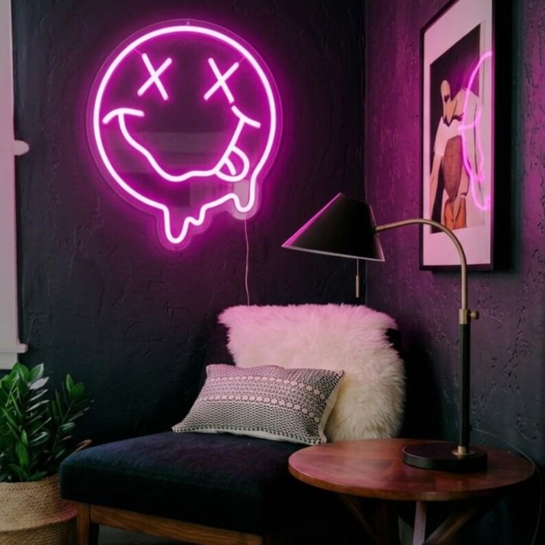 May include: A circular neon sign emitting a bright pink glow, featuring a stylized smiley face with X-shaped eyes and a tongue sticking out. The bottom edge of the circular face design appears to be melting with dripping neon light accents.
