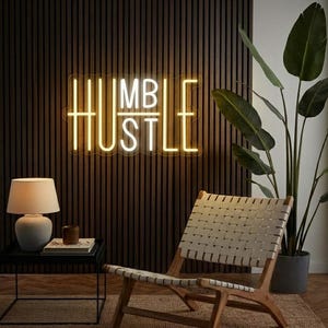 May include: A neon sign with the words "HUMBLE HUSTLE" in yellow and white against a dark wood-paneled wall. The room includes a woven chair, a table with a lamp, and a potted plant. The sign is a motivational piece of wall decor.