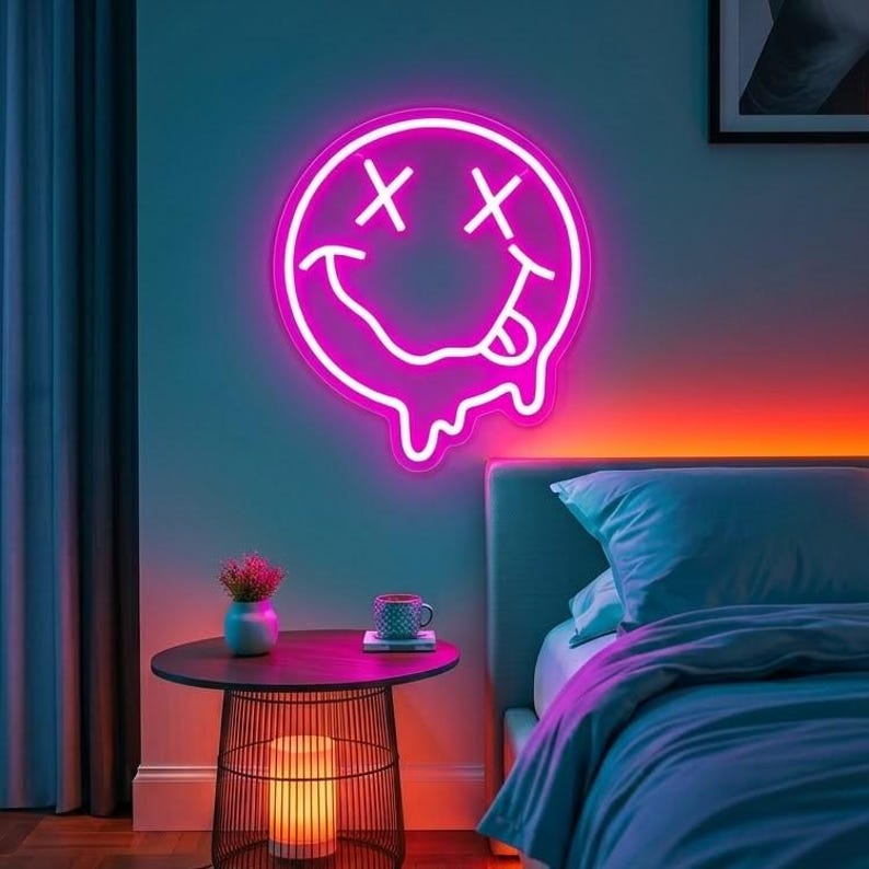 May include: A circular neon sign featuring a pink glowing smiley face with X-shaped eyes and a dripping mouth with a tongue sticking out. The sign is mounted on a wall above a round side table, emitting a vibrant pink light into the room.