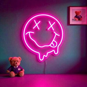 May include: A bright pink neon sign shaped like a circular smiley face with X-shaped eyes and a tongue sticking out, featuring a melting bottom edge. A brown teddy bear wearing a purple vest and glasses sits below the glowing neon wall light.