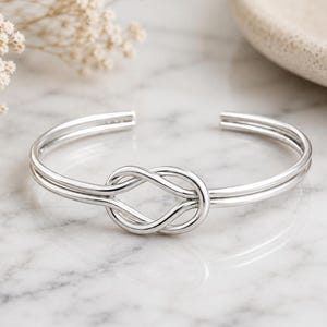 Könnte beinhalten: Ein silberfarbenes Armband in Manschettenform mit einem zentralen Unendlichkeitsknoten-Design. Das Armband besteht aus zwei parallelen Metallbändern, die in der Mitte zu einer symmetrischen Schlaufe zusammenlaufen und eine polierte Oberfläche bilden.