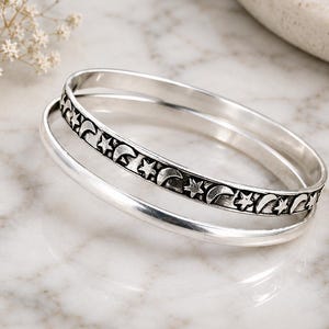 May include: Two silver-toned bangle bracelets. One bangle features a smooth, polished surface, while the other is engraved with a repeating pattern of crescent moons and five-pointed stars, finished with dark oxidation in the recessed areas.