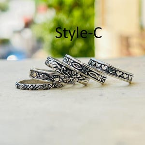 Könnte beinhalten: Eine Sammlung von vier Ringen aus silberfarbenem Metall mit der Aufschrift Style-C. Jeder Ring weist ein eigenes geprägtes Muster auf, darunter geometrische Formen, florale Motive und eine Reihe kleiner Herzen, alle mit oxidierter Optik.