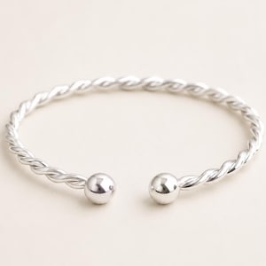 Könnte beinhalten: Ein silberfarbenes Armband in Manschettenform mit einem gedrehten, seilartigen Metallband. Das Armband hat ein offenes Design mit einer glatten, polierten kugelförmigen Perle an jedem der beiden Enden, die eine Kreisform mit einer Lücke bilden.