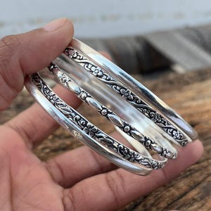 Könnte beinhalten: Ein Satz silberner Armreifen mit aufwendigen floralen und geometrischen Mustern. Die Armreifen sind gestapelt, wodurch die detaillierten Designs und die polierte Oberfläche hervorgehoben werden. Die Armbänder bestehen aus glänzendem Metall.