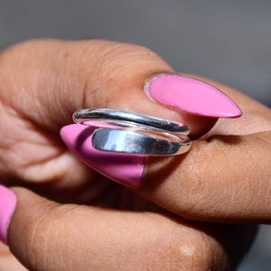 Könnte beinhalten: Ein polierter silberfarbener Bypass-Ring mit einer glatten, abgerundeten Ringschiene, die sich um den Finger legt. Der Ring hat eine reflektierende metallische Oberfläche mit einem einfachen, offenen Design, das sich leicht überlappt.