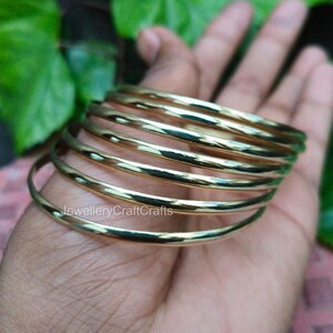 Könnte beinhalten: Ein Stapel goldfarbener Armreifen, jeder mit einem glatten, abgerundeten Design. Die Armreifen werden in einer Hand gehalten, mit grünem Laub im Hintergrund. Der Text "JewelleryCraftCrafts" ist auf einem der Armreifen zu sehen.
