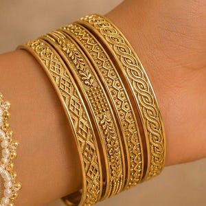 Könnte beinhalten: Eine Sammlung goldener Armreifen mit aufwendigen Mustern schmückt ein Handgelenk. Die Armreifen weisen detaillierte Designs auf, die eine Mischung aus Texturen zeigen. Das Bild hebt die Handwerkskunst und Eleganz des Schmucks hervor.