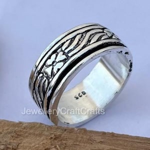 Könnte beinhalten: Ein Silberring mit breiter Ringschiene, die ein geprägtes Blumen- und Flechtmuster aufweist. Die Innenseite des Rings ist mit der Zahl 925 gestempelt, was auf Sterlingsilber hinweist. Der Ring hat eine polierte Oberfläche mit dunklen Vertiefungen.