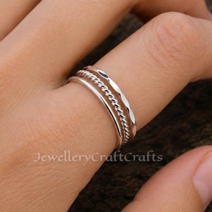 Könnte beinhalten: Ein Set aus drei silbernen Ringen, jeder mit einem einzigartigen Design. Ein Ring ist glatt, einer hat eine gedrehte Seiltextur und der dritte ein facettiertes Muster. Die Ringe sind auf einem Finger gestapelt.