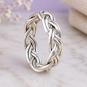 Könnte beinhalten: Ein silberner Ring mit einem durchgehenden geflochtenen oder gewebten Metalldesign. Das Band besteht aus mehreren Strängen aus poliertem Silber, die ineinandergreifen und ein strukturiertes, kreisförmiges Muster um den Ring bilden.