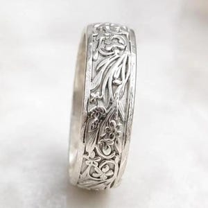 May include: A silver band ring featuring an intricate, embossed floral and vine pattern covering the entire outer surface. The metal has a polished finish, highlighting the detailed botanical engravings that wrap around the circumference of the ring.