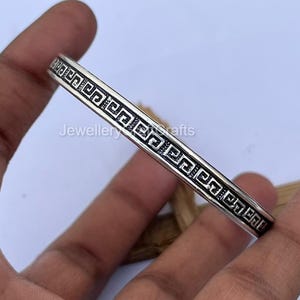 Könnte beinhalten: Ein silberfarbenes Armreif-Armband mit einem sich wiederholenden geometrischen Muster. Das Armband wird in einer Hand gehalten, was das aufwendige Design zeigt. Das Schmuckstück ist ein stilvolles Accessoire.