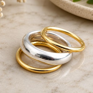 May include: A set of three stacked rings consisting of two thin gold-toned bands and one thicker silver-toned band. The rings have a smooth, polished finish and are arranged in a cluster, showcasing a combination of metallic gold and silver colours.