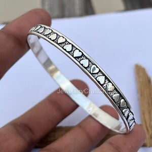 Könnte beinhalten: Ein silbernes Armreif-Armband mit einem sich wiederholenden Herzmuster. Die Herzen sind gleichmäßig um das Armband herum angeordnet. Das Armband hat eine glatte, glänzende Oberfläche und reflektiert das Licht.