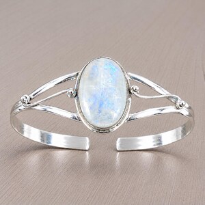 Könnte beinhalten: Ein silbernes Armband mit einem großen, ovalen Mondstein-Cabochon in der Mitte. Das Metallband hat eine polierte Oberfläche mit dekorativen, geschwungenen Drahtakzenten auf beiden Seiten des Steins, was ein offenes Design mit geteilter Schiene erzeugt.