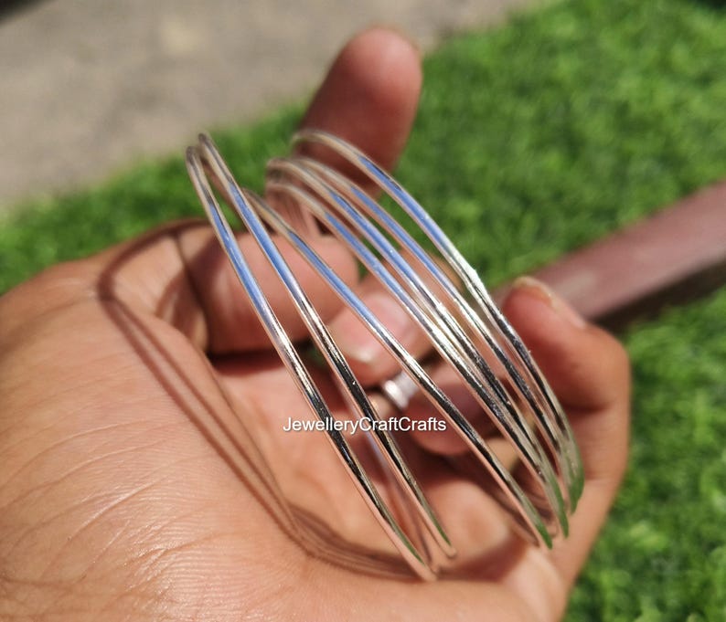 Peut inclure: Plusieurs bracelets argent&eacute;s, fins et lisses, sont tenus dans une main. Les bracelets brillent et refl&egrave;tent la lumi&egrave;re. L'image est prise &agrave; l'ext&eacute;rieur, avec un fond vert. Le texte "JewelleryCraftCrafts" est visible.
