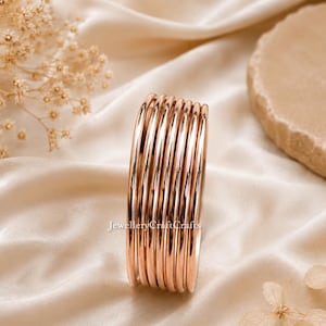 Könnte beinhalten: Ein Stapel roségoldener Armreifen. Die glatten, glänzenden Armbänder sind aufeinander gestapelt und reflektieren das Licht. Der Schmuck wird auf einem cremefarbenen Satinstoff präsentiert. Der Hintergrund umfasst getrocknete Blumen und einen Stein.