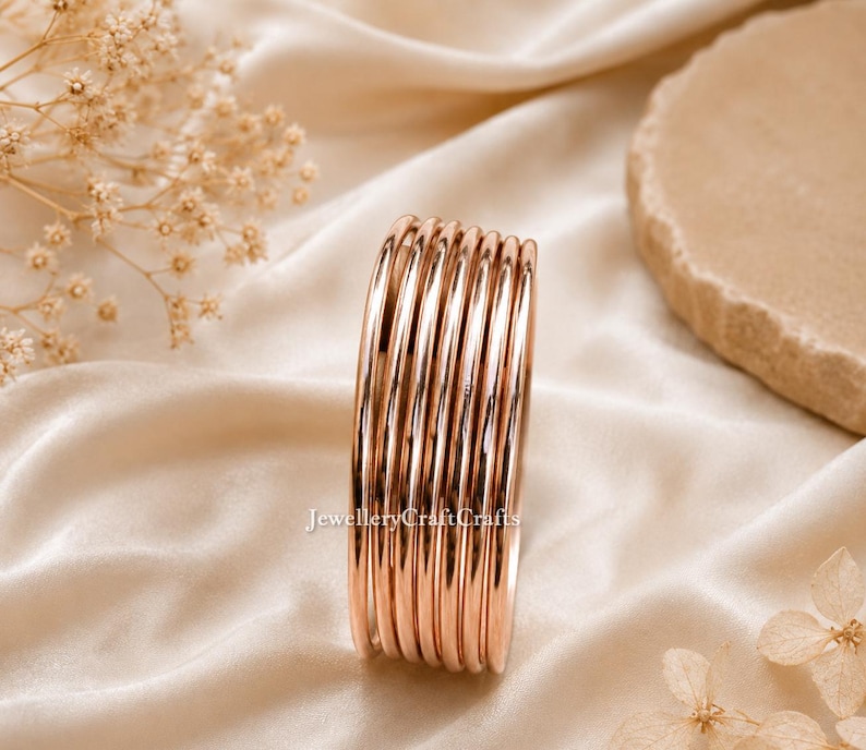 May include: A collection of rose gold bangles. The smooth, shiny bracelets are stacked together, reflecting light. The jewellery is displayed on a cream-coloured satin fabric. The background includes dried flowers and a stone.