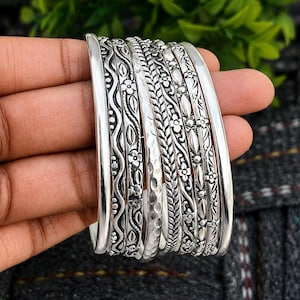 May include: A set of silver bangles with detailed floral and geometric designs. The bangles showcase embossed flowers, leaves, and textured elements. The jewellery is presented against a blurred green and grey backdrop, highlighting the intricate patterns.