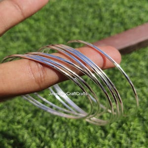 Peut inclure: Plusieurs bracelets fins en argent. Les bracelets sont empil&eacute;s, refl&eacute;tant la lumi&egrave;re. L'image est prise &agrave; l'ext&eacute;rieur avec un fond vert. Les bracelets sont tenus par les doigts d'une personne.