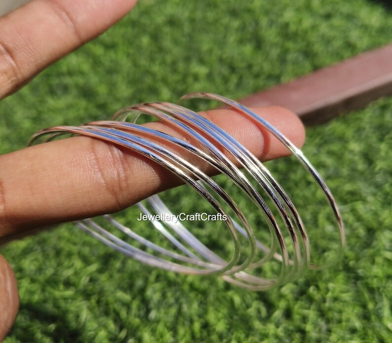 Peut inclure: Plusieurs bracelets fins en argent. Les bracelets sont empil&eacute;s, refl&eacute;tant la lumi&egrave;re. L'image est prise &agrave; l'ext&eacute;rieur avec un fond vert. Les bracelets sont tenus par les doigts d'une personne.