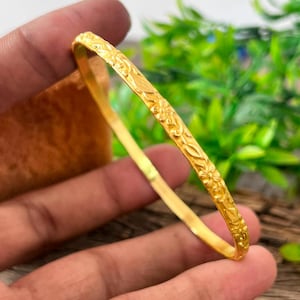 Könnte beinhalten: Ein dünner, kreisförmiger Goldarmreif mit einem durchgehenden, geprägten Blumenmuster auf der gesamten Außenfläche. Das Metall hat ein helles, poliertes Gelbgold-Finish, das das Licht auf den detaillierten, erhabenen Blumenmotiven reflektiert.
